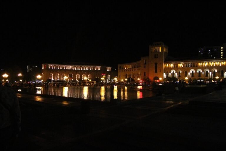 Buildings in a city square lit up at night, with their reflections in the water.